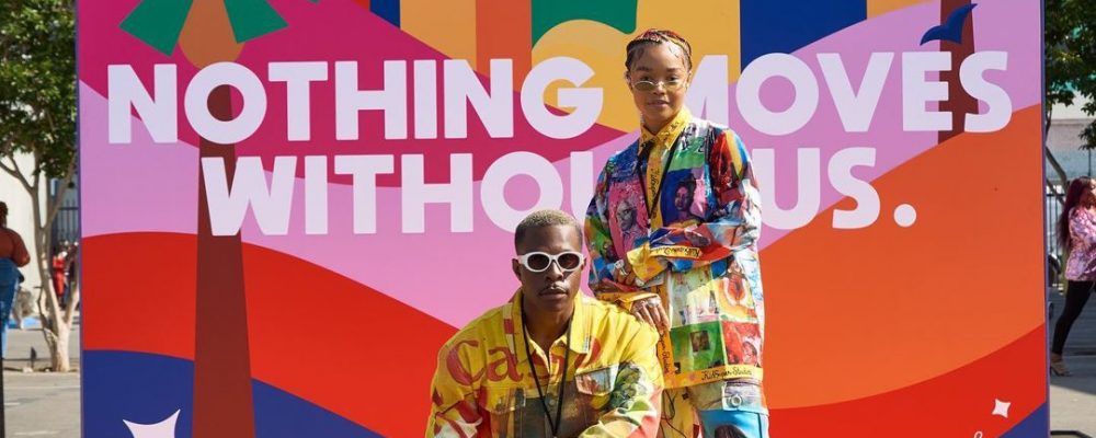 TWO PEOPLE POSE IN FRONT OF “NOTHING MOVES WITHOUT US.” WRITTEN ON COLORFUL BACKDROP AT CULTURECON NYC EVENT